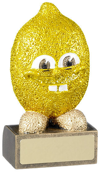 Bright golden glittery lemon-shaped trophy with large googly eyes, smiling mouth showing white teeth, textured surface, standing on dark base with engraving plate