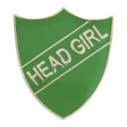 Green shield-shaped enamel pin badge with gold border showing white 'HEAD GIRL' text on diagonal banner design against white background